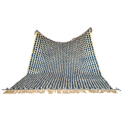 This image showcases a handmade Moroccan wool rug featuring a bold deep blue geometric pattern against a natural beige background. The rug is rectangular with hand-knotted fringe on both ends, giving it a bohemian and artisanal touch. Perfect for modern or traditional interiors, the rug is placed in a minimalist living room with earthy tones and natural light, highlighting its soft texture and intricate craftsmanship.